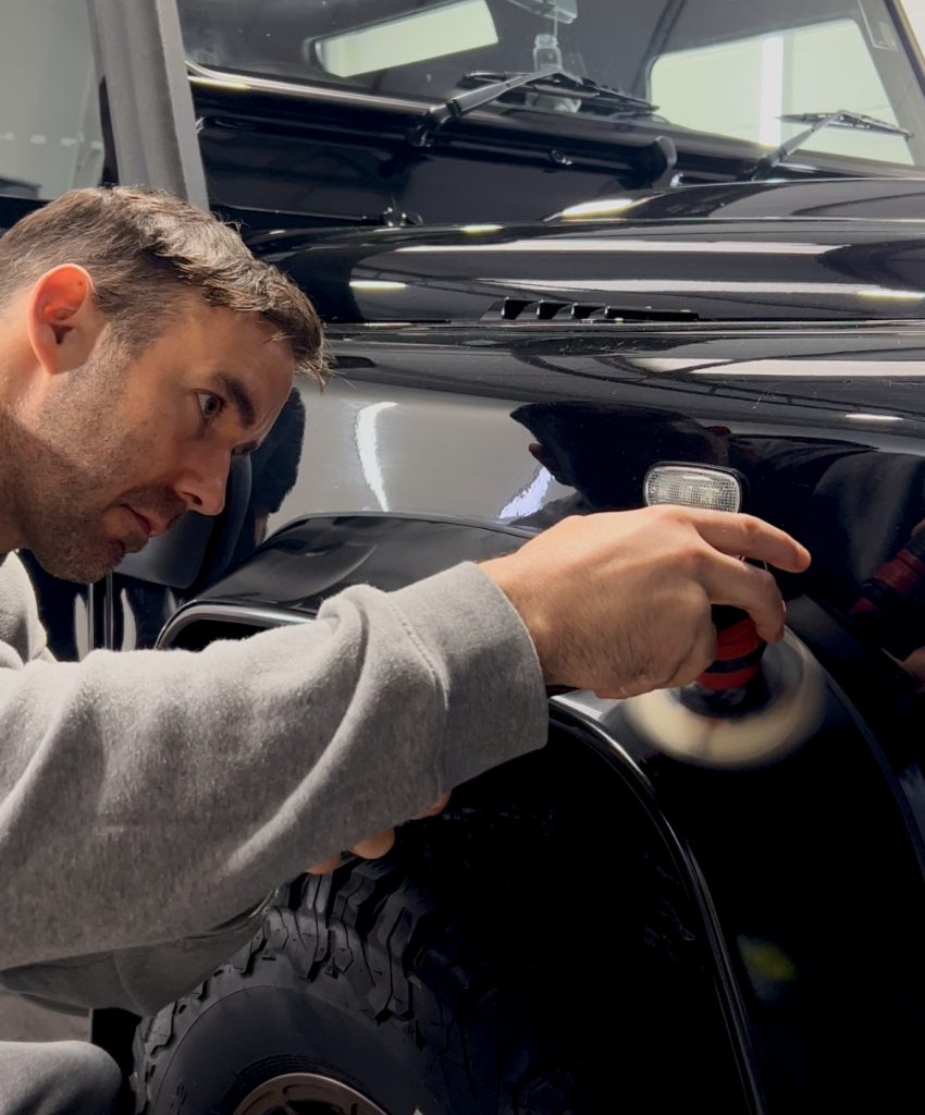 Male wearing grey jumper polishing a black car with handheld polisher